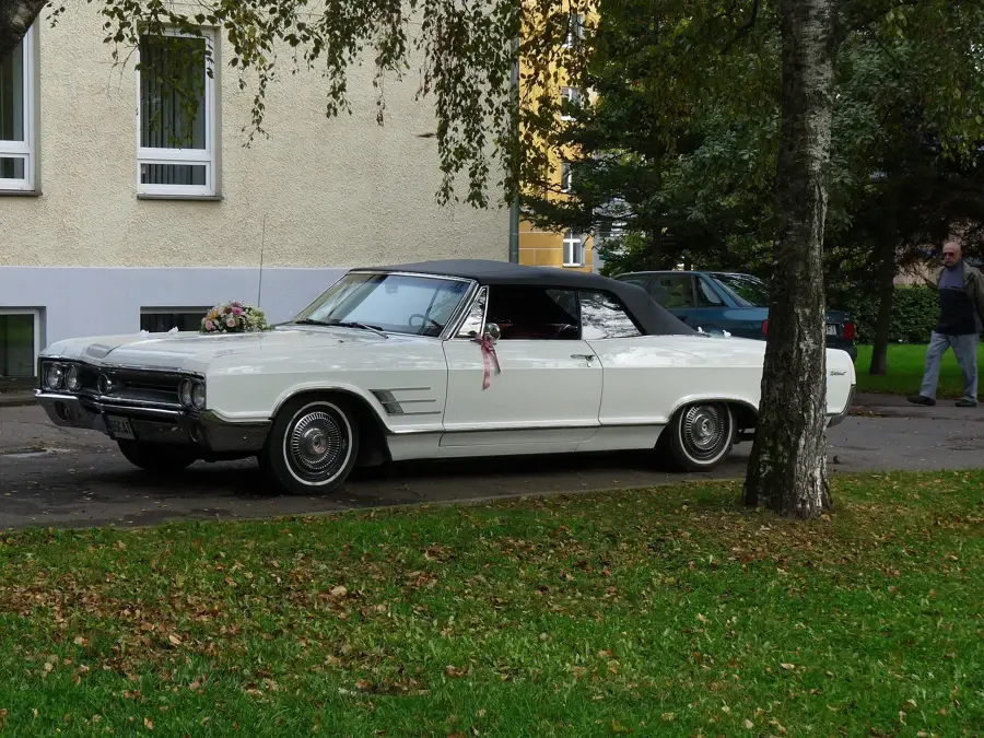 Voiture de mariés blanche décorée d'un bouquet sur le capot et d'un ruban rose